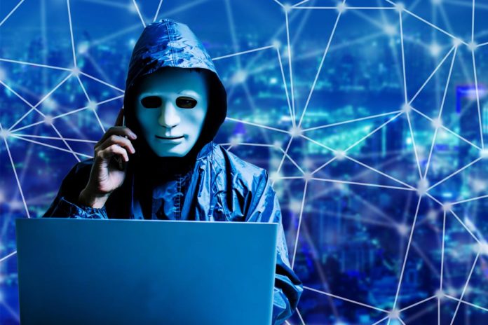 Anonymous computer hacker in white mask and hoodie. Obscured dark face using laptop computer for cyber attack and calling on cellphone on modern city background, Data thief, internet attack, darknet and cyber security concept.